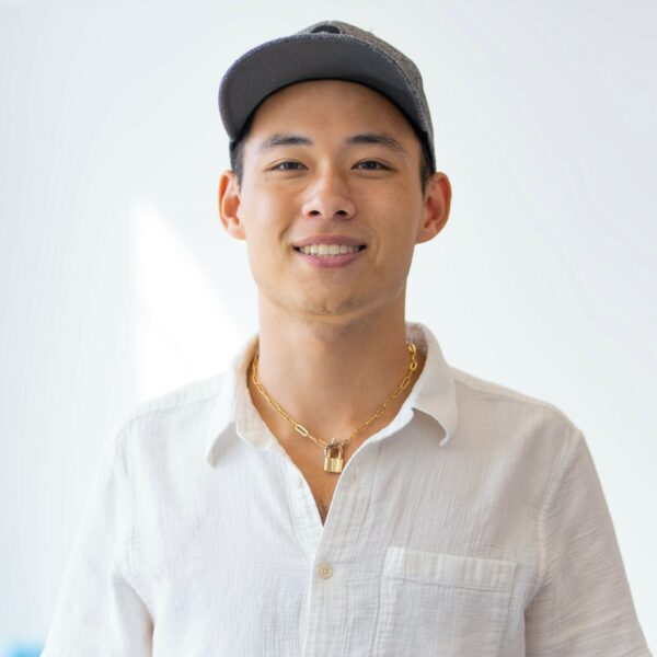 A smiling man wearing a cap and a necklace, dressed in a casual buttoned shirt.
