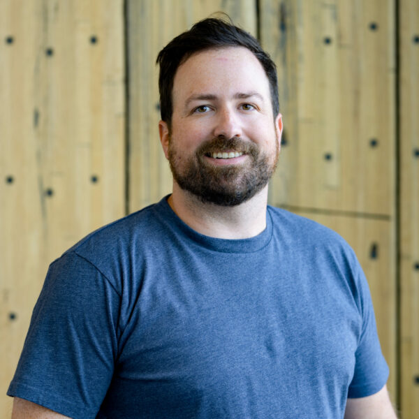 A smiling man with a beard, wearing a blue t-shirt, standing in front of a wooden background.