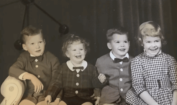 Four young children, three boys in buttoned shirts and bow ties and one girl in a checkered dress, sit together and smile for a black-and-white portrait.