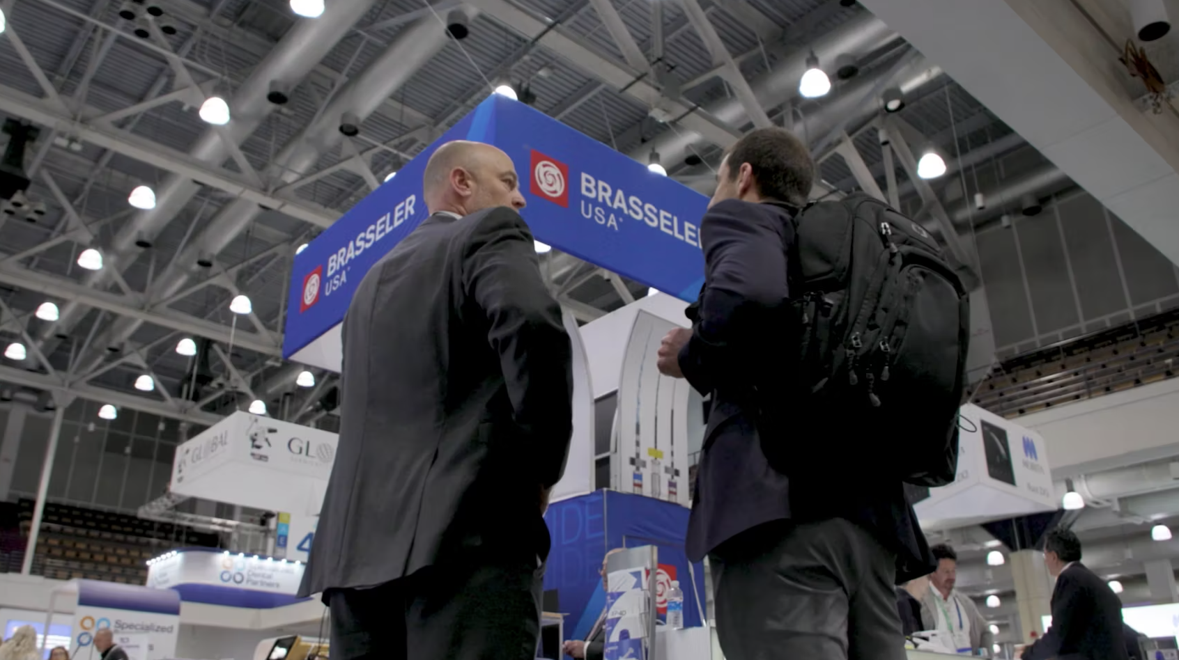 Two men in business attire stand under a Brasseler USA booth at a convention or trade show, engaged in conversation. Industrial lighting and other booths are visible in the background.