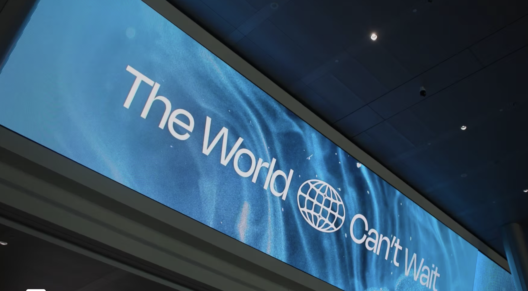 Large digital sign with blue background displays the words “The World Can’t Wait” alongside a globe icon, viewed from below inside a modern building.