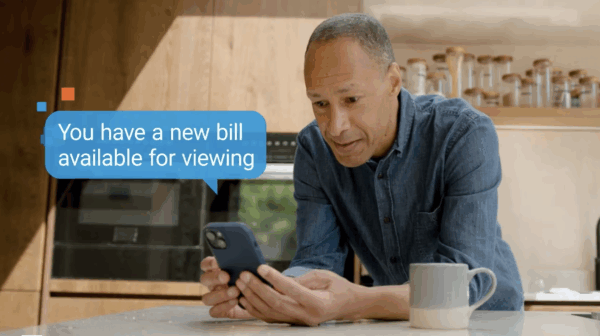 A man in a kitchen looks at his phone, which displays a notification bubble: "You have a new bill available for viewing." A mug sits on the counter beside him.