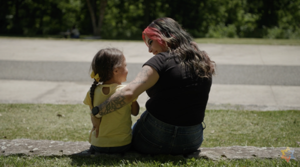 An adult and a child sit on a grassy area facing away, with the adult's arm around the child. Both have dark hair and wear casual clothes; the child has a yellow bow.