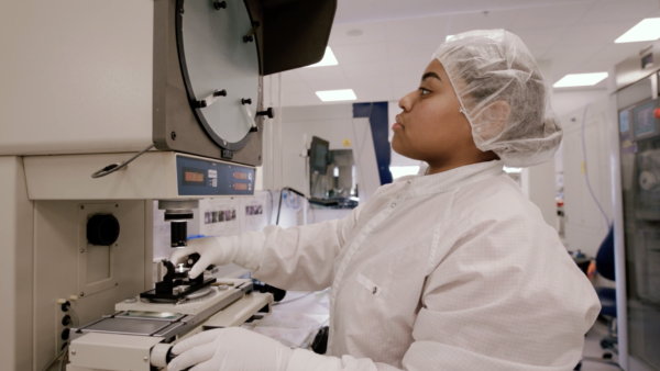 A person in a lab coat, hairnet, and gloves operates scientific equipment in a laboratory setting.