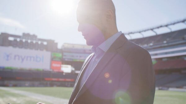 A person in a suit stands on a sports field with sunlight and lens flare partially obscuring their face. Stadium seats and a large screen are visible in the background.