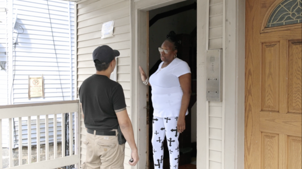 A person stands at the doorway talking to a woman who is standing just inside her front door of a beige house.