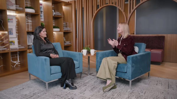 Two women sit in blue armchairs facing each other in a modern room with bookshelves, a small table with mugs between them, having a conversation.