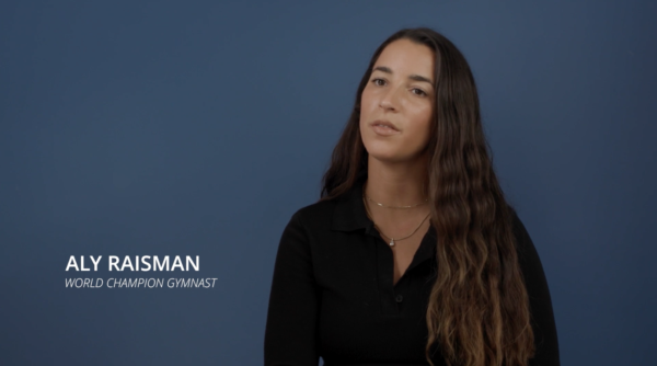 A woman with long wavy brown hair wearing a black top sits against a plain blue background. Text on the image reads: "Aly Raisman, World Champion Gymnast.