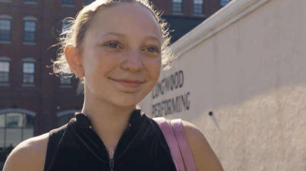 A young person with light hair tied back, wearing a black zip-up top and pink bag strap, stands outside in sunlight near a light-colored wall with partially visible text.