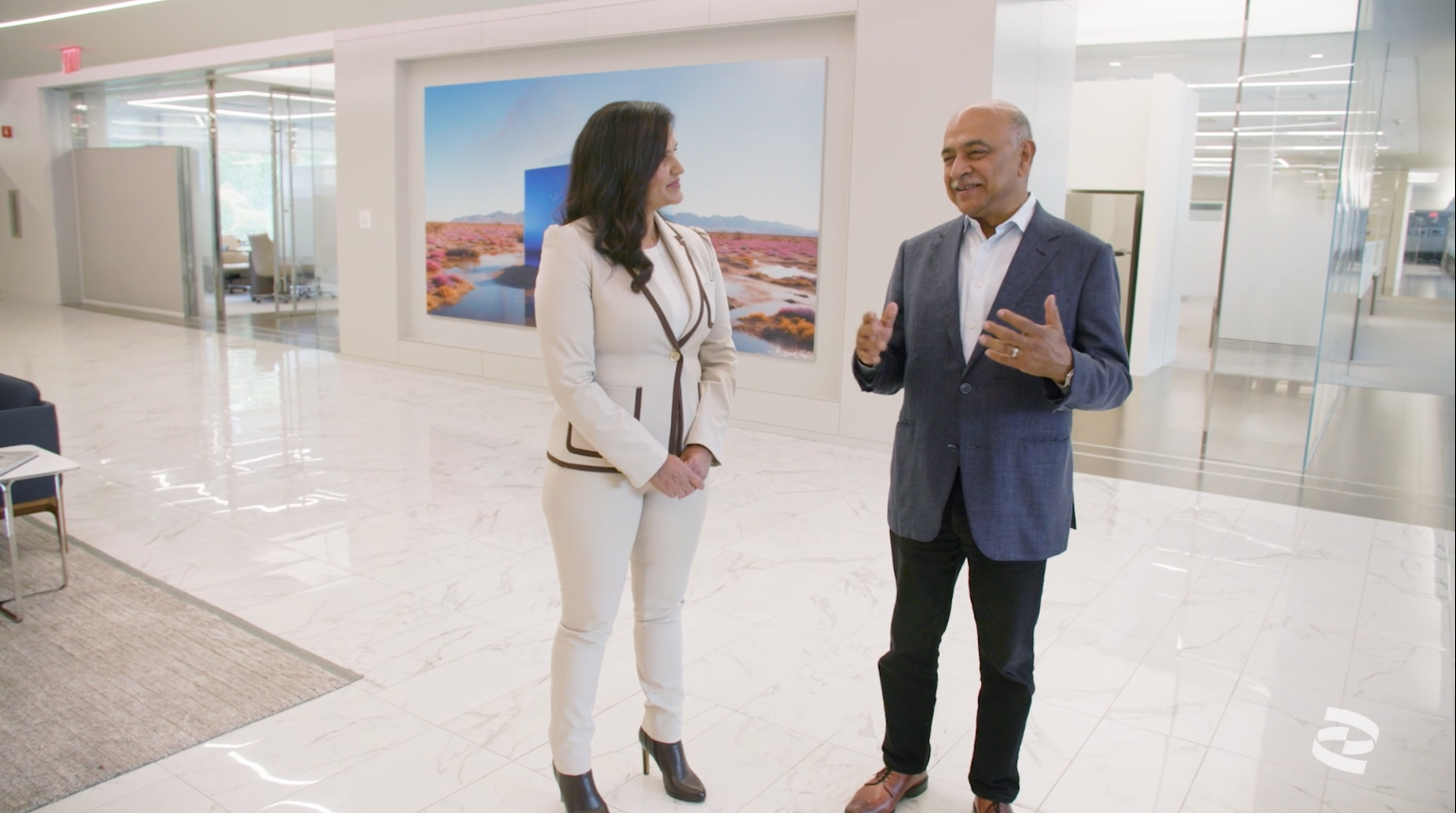 Two people stand and talk in a modern office lobby with white marble floors and a large landscape photo on the wall behind them.