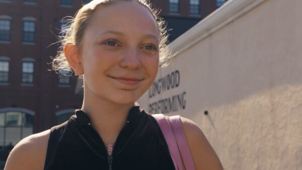 A young person with light hair pulled back smiles outdoors, wearing a black zip-up top and a pink bag. A beige building and a red brick building complete the scene—captured by a Boston Video Production Company.