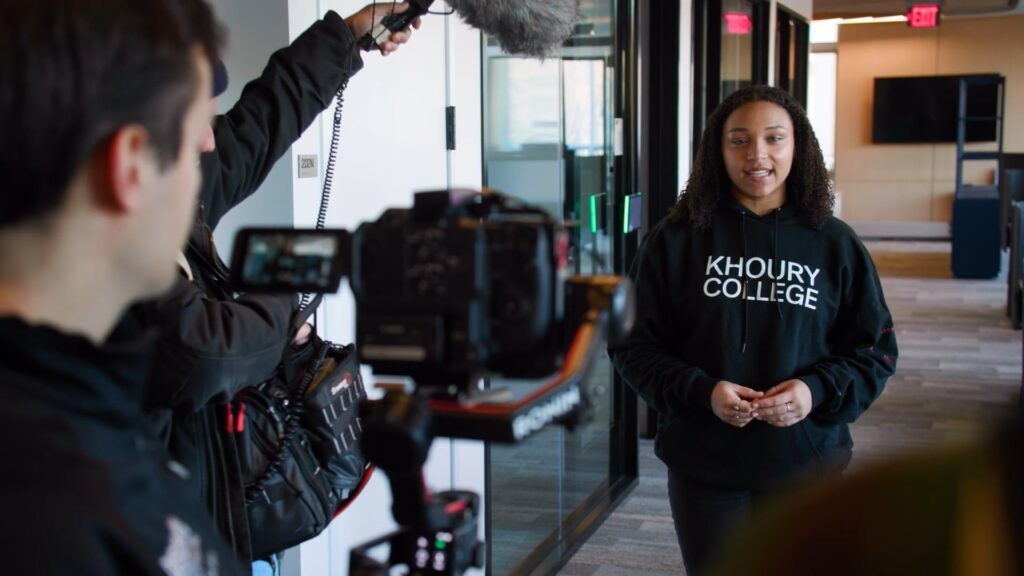p8 A woman in a "Khoury College" hoodie speaks in front of a camera and microphone setup in a hallway while being filmed by a crew, discussing the impact of technology across various industries.