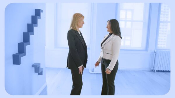 Two women in business attire stand facing each other and talking in a bright, modern office with geometric wall art and large windows—an ideal setting for a Boston Video Production Company shoot.